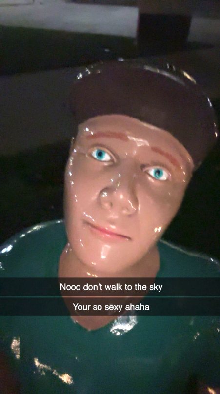 A photo taken with a smartphone camera at night of one of the sculptures of a person at the base of walking to the sky, with harsh front-camera selfie lighting. A Snapchat-style text overlay reads: "Nooo don't walk to the sky, your [sic] so sexy ahaha"